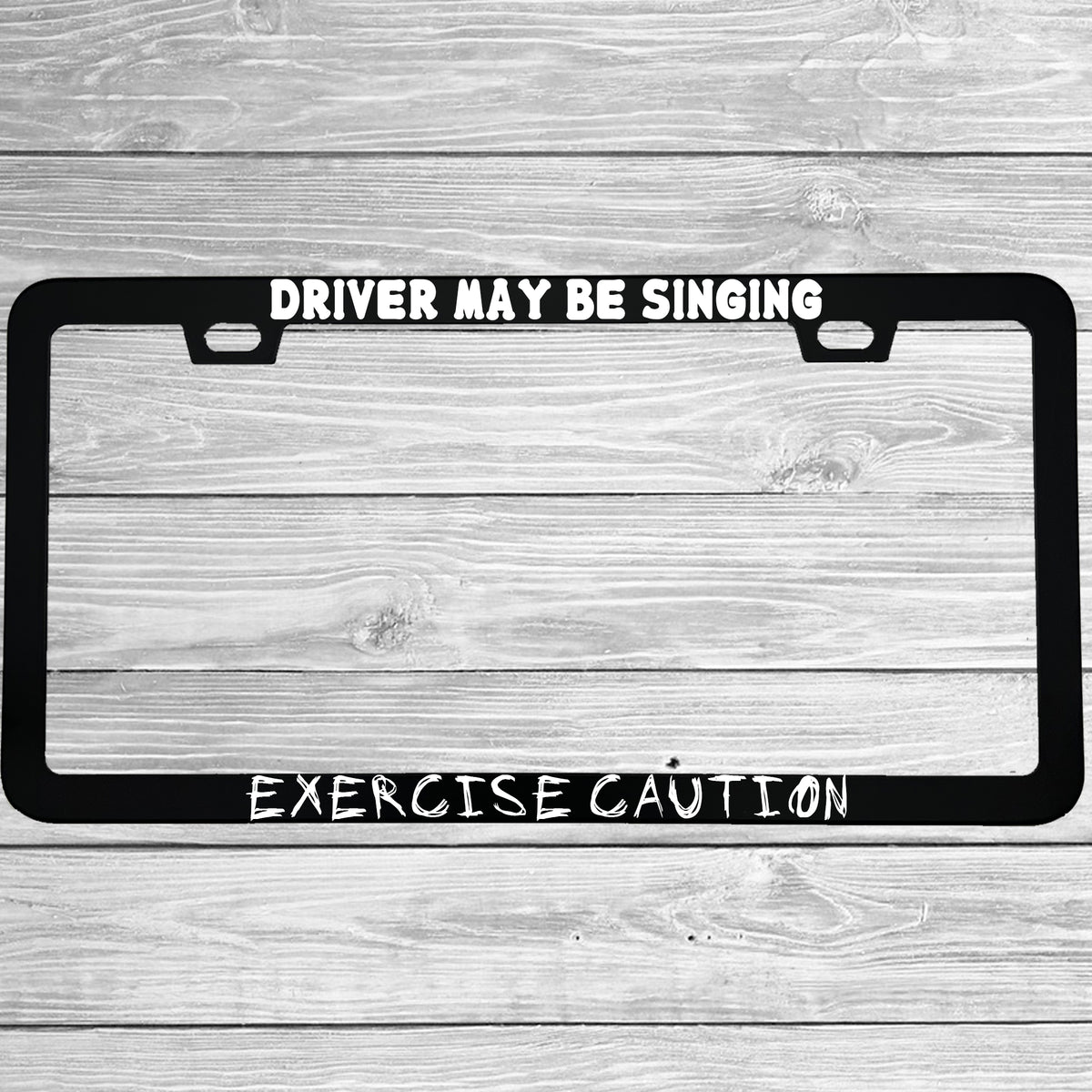 Close-up of "Driver May Be Singing Exercise Caution" license plate frame showing fade-proof print quality