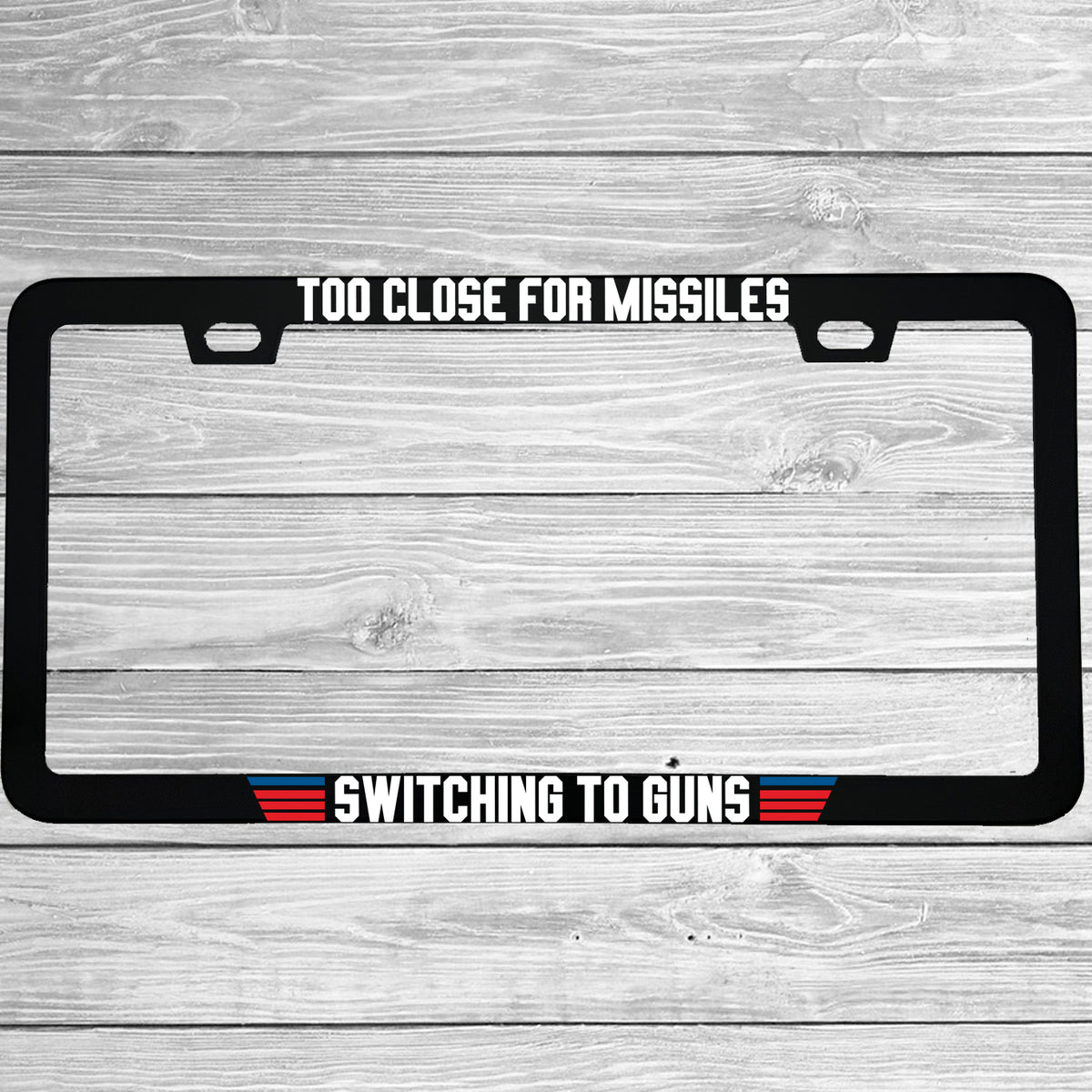 Close-up of "Too Close For Missiles Switching To Guns" license plate frame showing fade-proof print quality